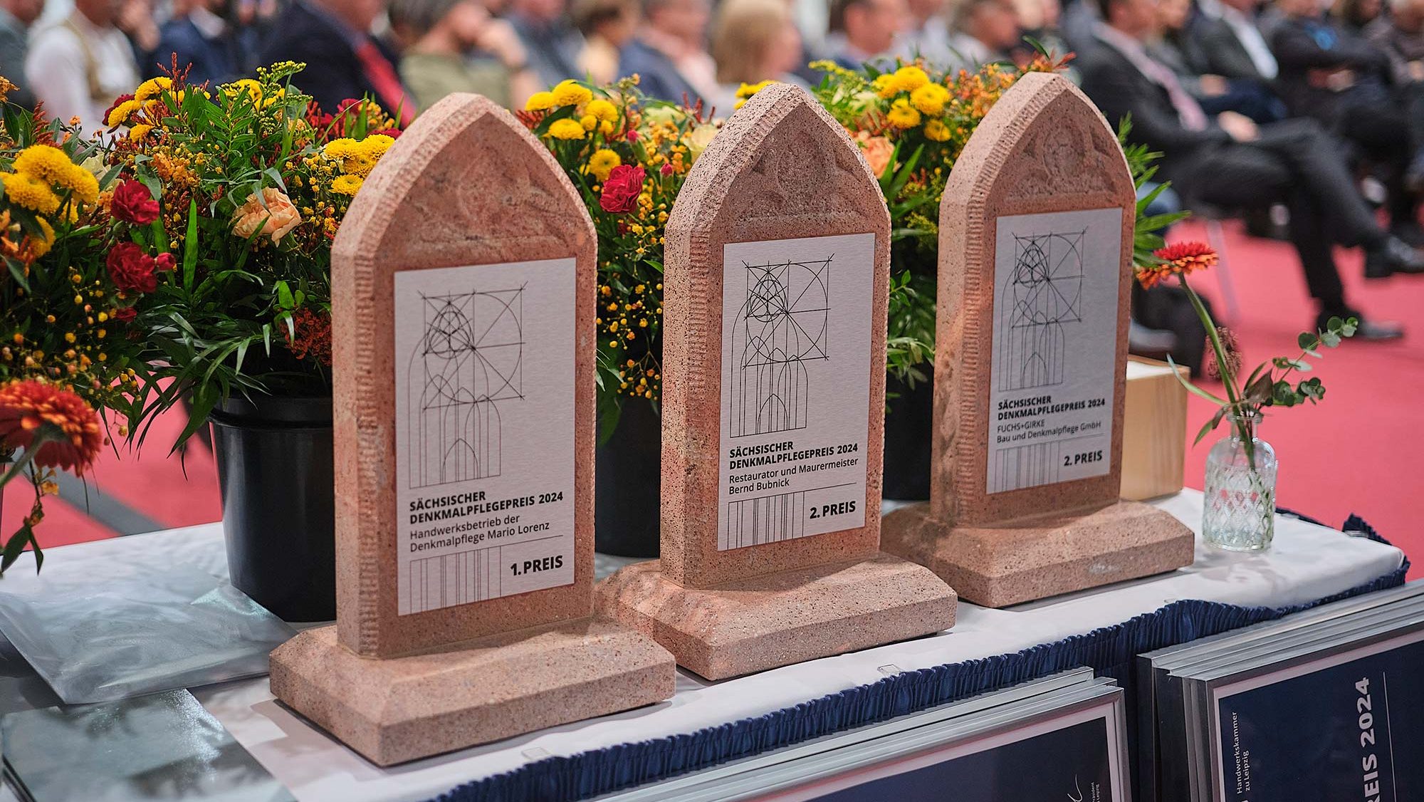 Sächsischer Denkmalpflegepreis - die drei Stelen der Preisträger vor Blumensträußen, Publikum in Hintergrund
